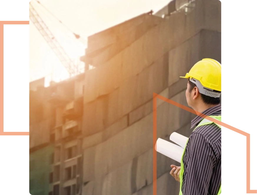 A person in a hard hat and safety vest holds blueprints at a commercial construction site, illuminated by the warm glow of sunrise or sunset.